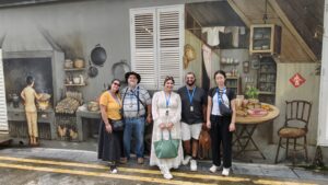 Walking tour participants pose in front of a detailed heritage mural depicting a traditional kitchen scene, complete with a wood-fired stove, hanging pots, and a table set with festive foods.